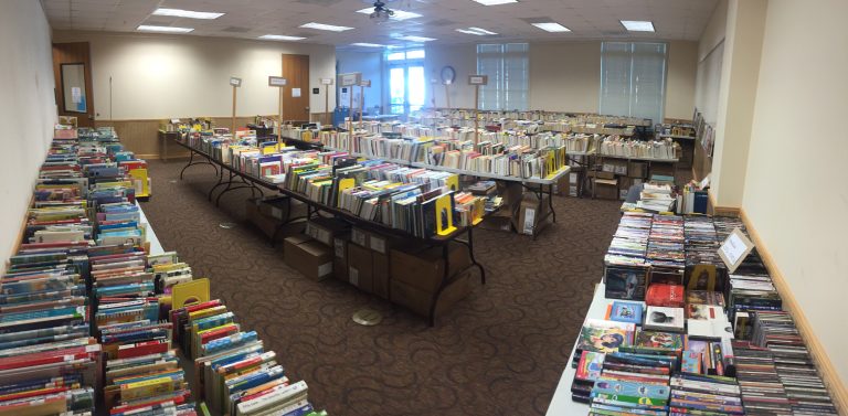 Long tables of donated books, sorted by category and labeled, filling the library meeting room before a Friends book sale.