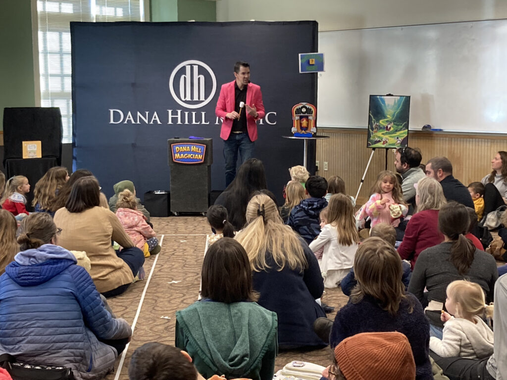 A library meeting room packed with children and parents sitting on the floor for a magic show.