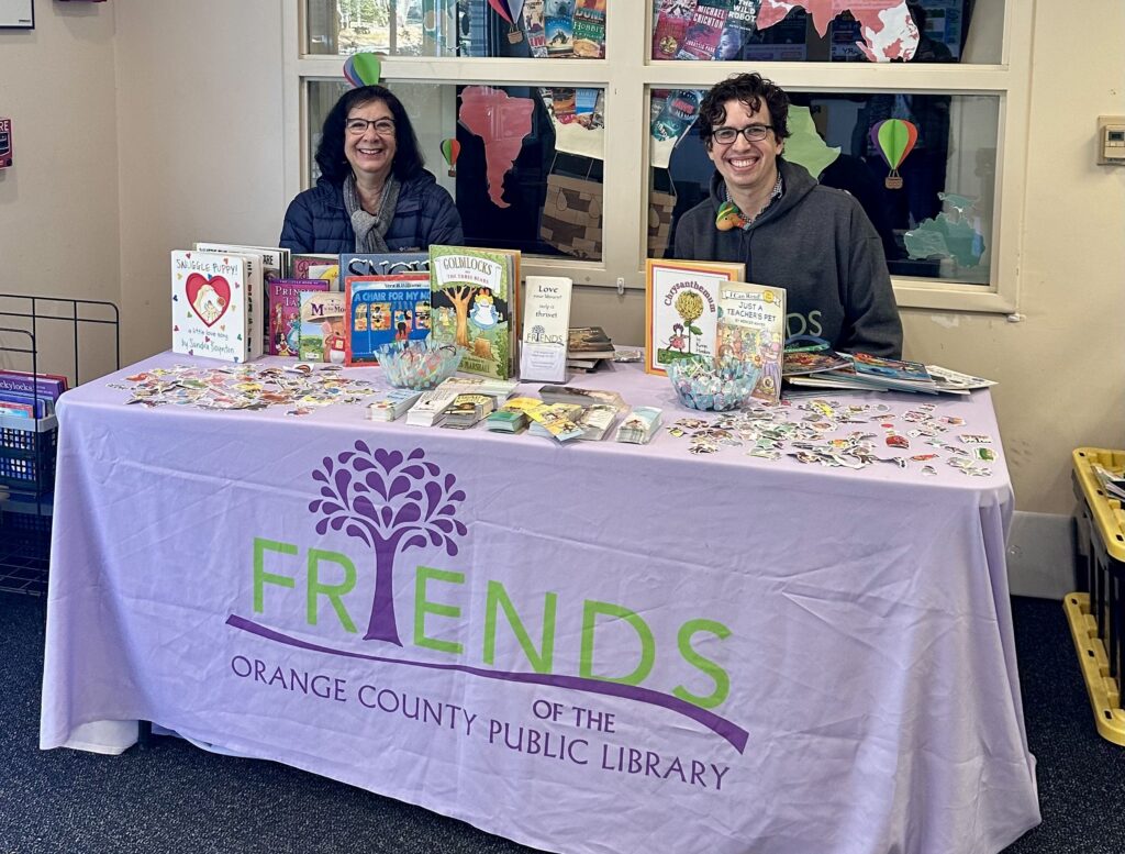 Two Friends volunteers behind a Friends of the Orange County Public Library table, handing out childrens books.