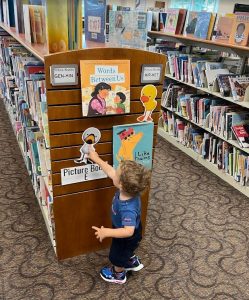 A young child reaching for a book on a picture-book display stand between the library shelves.
