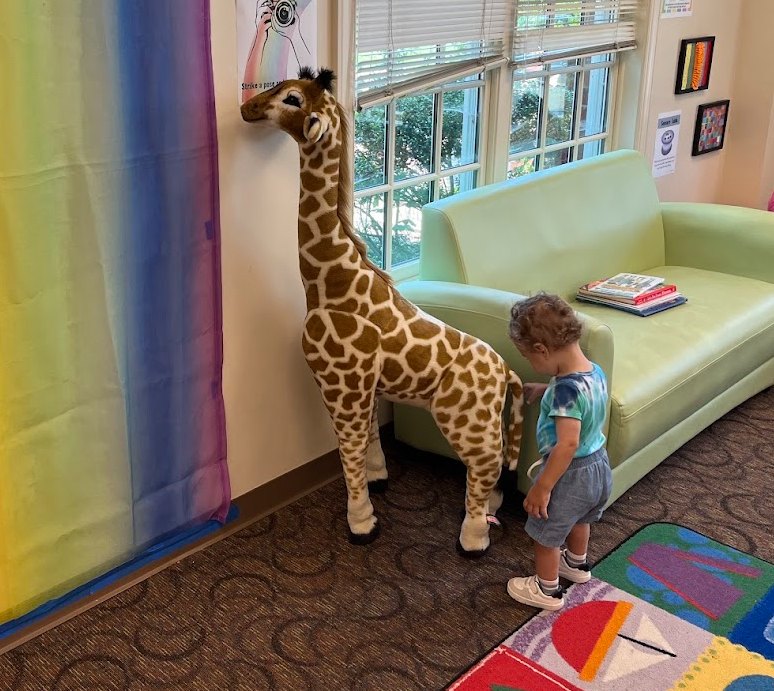 A toddler standing beside a life-size stuffed giraffe in the library's children's corner, next to a green couch stacked with picture books.