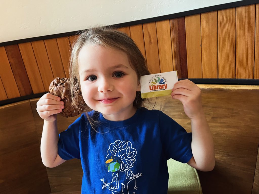 A smiling young child holding up an Orange County Public Library card in one hand and a chocolate cookie in the other
